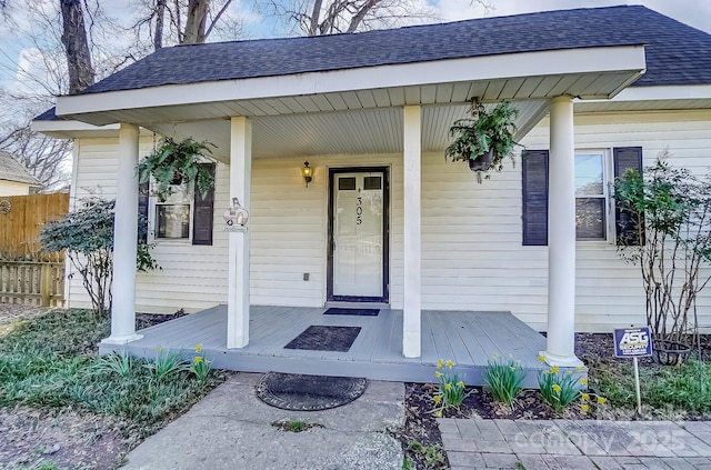 view of exterior entry featuring a porch, roof with shingles, and fence