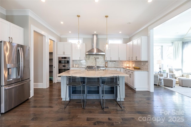 kitchen featuring an island with sink, appliances with stainless steel finishes, white cabinets, wall chimney exhaust hood, and pendant lighting