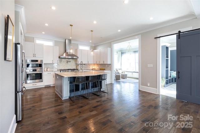 kitchen featuring white cabinets, wall chimney range hood, decorative light fixtures, a barn door, and a kitchen island with sink