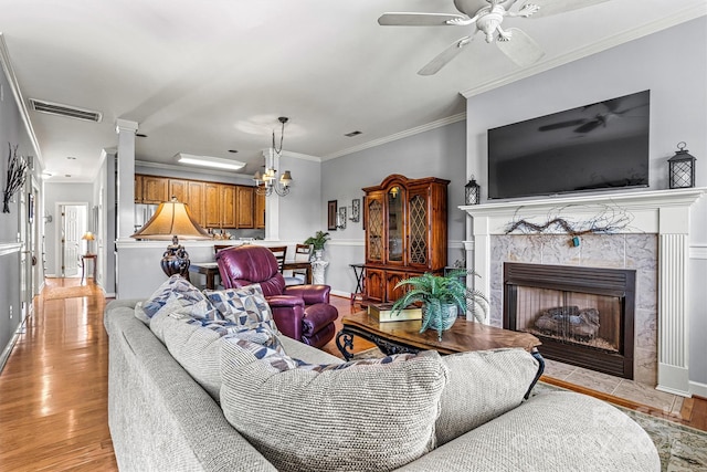 living room featuring light wood finished floors, visible vents, a high end fireplace, ornamental molding, and ceiling fan with notable chandelier