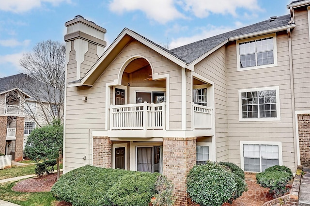 townhome / multi-family property featuring a balcony, roof with shingles, a chimney, and brick siding