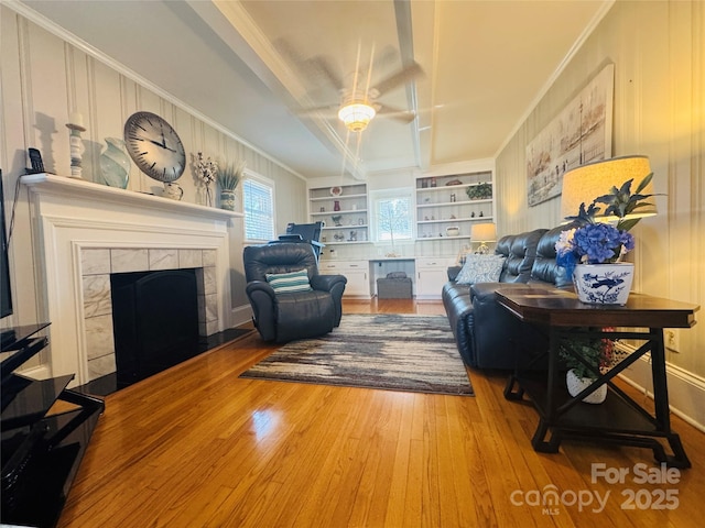 living room with crown molding, built in features, ceiling fan, a tiled fireplace, and hardwood / wood-style floors