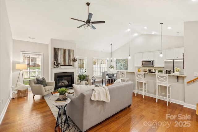 living room featuring baseboards, a ceiling fan, a glass covered fireplace, light wood-style flooring, and high vaulted ceiling