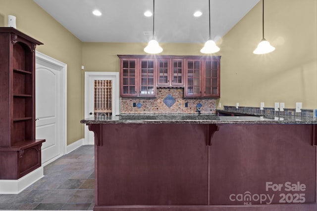 kitchen featuring decorative light fixtures, a peninsula, backsplash, dark stone countertops, and glass insert cabinets