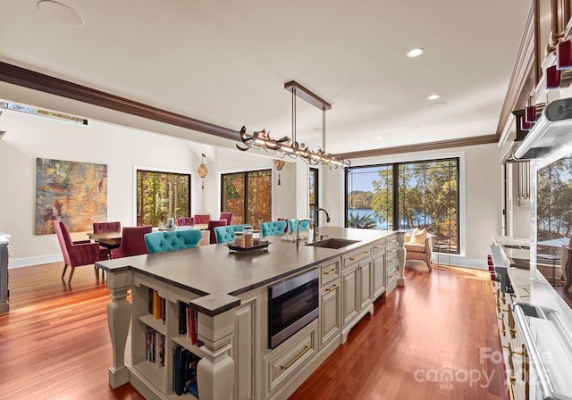 kitchen with a center island with sink, sink, stainless steel microwave, crown molding, and hanging light fixtures