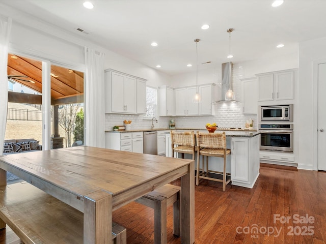 kitchen with dark wood-style floors, a kitchen island, appliances with stainless steel finishes, wall chimney exhaust hood, and backsplash