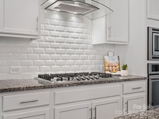 kitchen featuring decorative backsplash, range hood, appliances with stainless steel finishes, dark stone countertops, and white cabinets