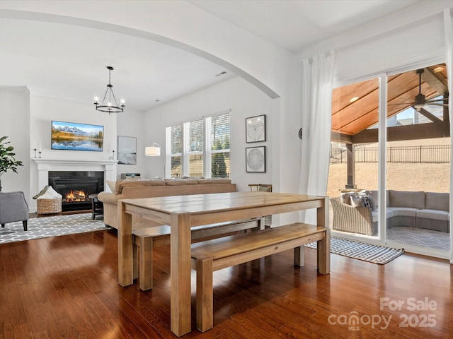 dining room with a glass covered fireplace, ceiling fan with notable chandelier, dark wood finished floors, and arched walkways