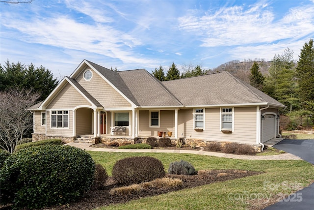 view of front of home featuring an attached garage, covered porch, driveway, roof with shingles, and a front yard