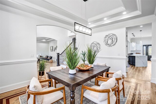 dining room with arched walkways, light wood-style flooring, stairway, a raised ceiling, and crown molding