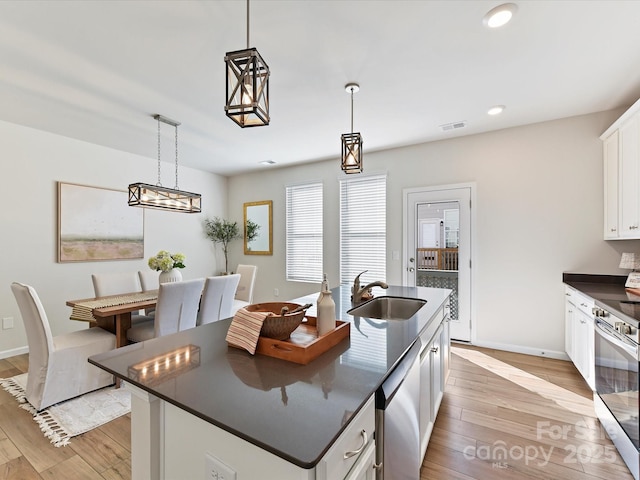 kitchen with decorative light fixtures, dark countertops, white cabinets, a sink, and an island with sink