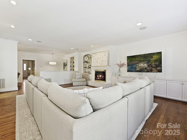 living area featuring a warm lit fireplace, built in features, dark wood-style floors, crown molding, and recessed lighting