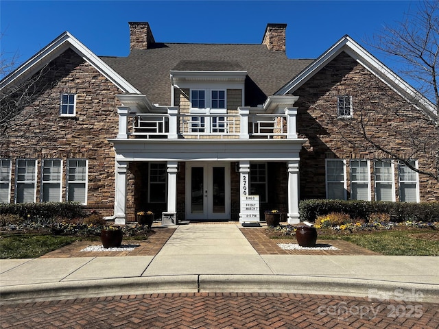 view of front of house featuring french doors, stone siding, a balcony, and a chimney