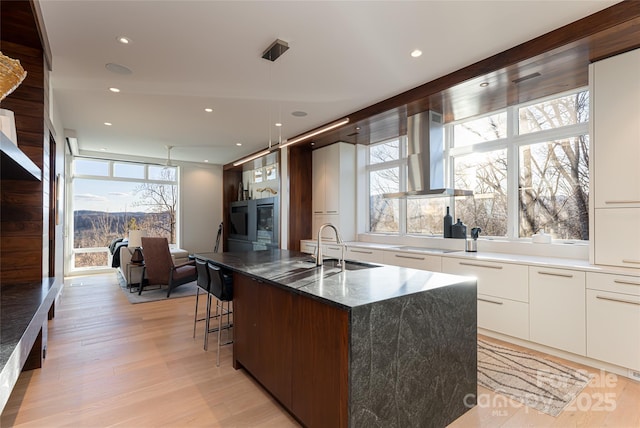 kitchen featuring white cabinets, light wood-style floors, wall chimney range hood, modern cabinets, and a center island with sink