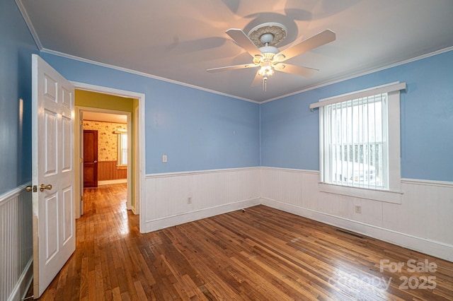 empty room with visible vents, a ceiling fan, ornamental molding, wainscoting, and hardwood / wood-style floors