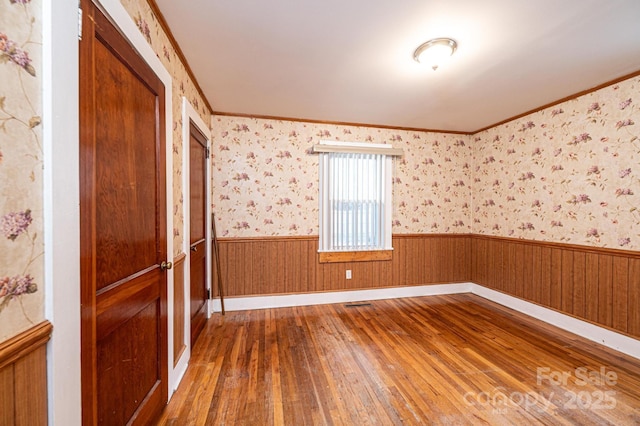 empty room with crown molding, wainscoting, wood-type flooring, and wallpapered walls