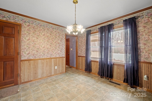 spare room featuring crown molding, visible vents, an inviting chandelier, wainscoting, and wallpapered walls