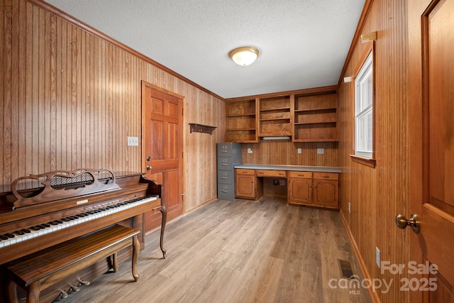 office featuring light wood finished floors, wooden walls, ornamental molding, a textured ceiling, and built in desk