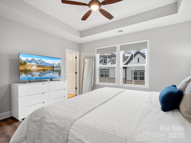 bedroom with ceiling fan, visible vents, baseboards, dark wood-style floors, and a tray ceiling