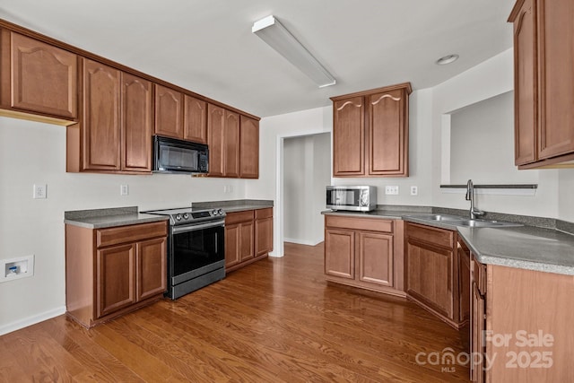 kitchen with dark countertops, appliances with stainless steel finishes, brown cabinetry, dark wood-type flooring, and a sink