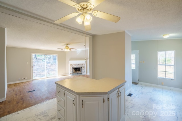 kitchen with white cabinetry, a center island, a stone fireplace, ceiling fan, and a textured ceiling