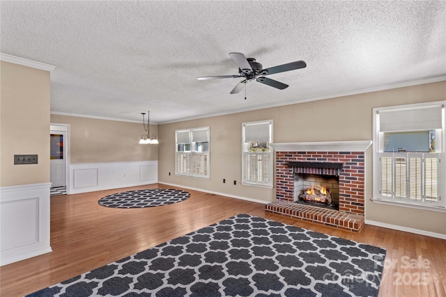 living room with ceiling fan with notable chandelier, wood finished floors, wainscoting, a brick fireplace, and crown molding