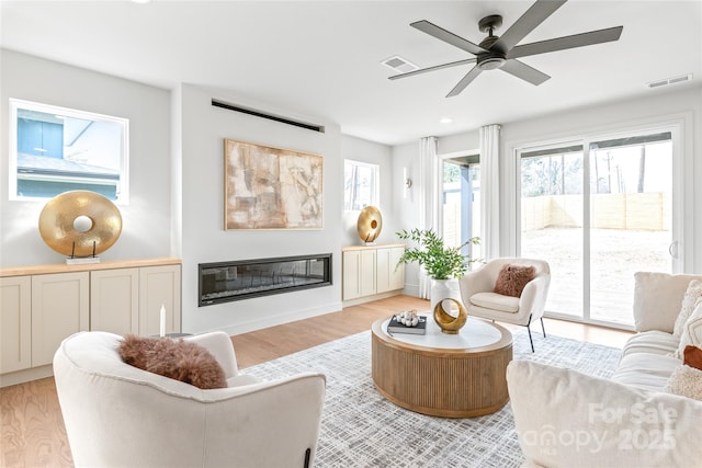 interior space featuring ceiling fan, light wood-type flooring, a glass covered fireplace, and visible vents