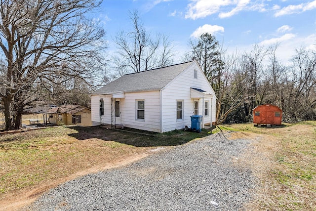 bungalow-style house featuring an outbuilding, gravel driveway, a storage unit, a shingled roof, and a front lawn