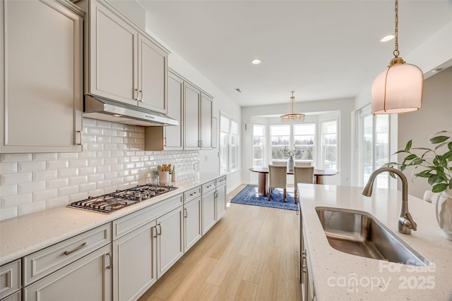 kitchen with stainless steel gas stovetop, light wood-style flooring, decorative backsplash, a sink, and under cabinet range hood