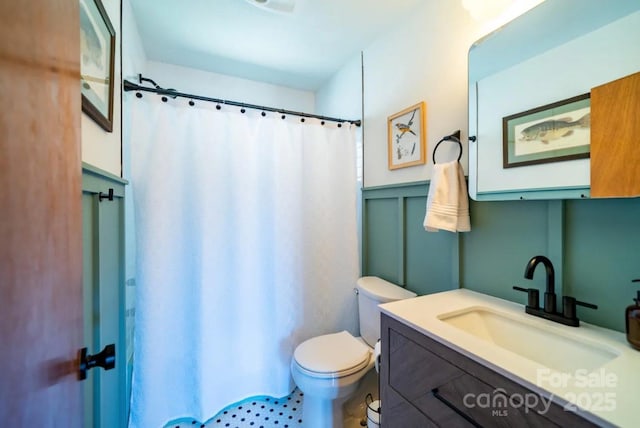 full bathroom featuring vanity, a shower with curtain, toilet, wainscoting, and a decorative wall