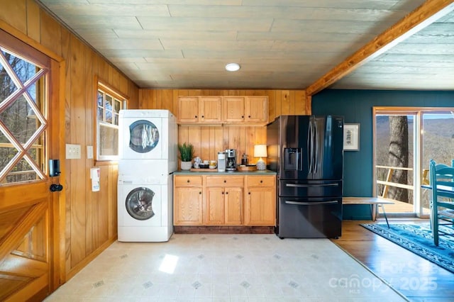 kitchen featuring stacked washing maching and dryer, wooden walls, plenty of natural light, and black fridge with ice dispenser