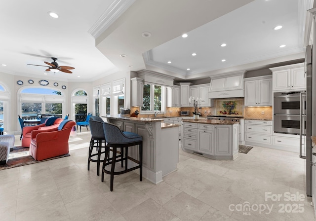 kitchen with double oven, stone countertops, a kitchen island with sink, premium range hood, and a breakfast bar