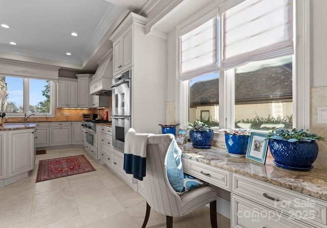 kitchen featuring premium range hood, white cabinetry, light stone countertops, built in desk, and crown molding