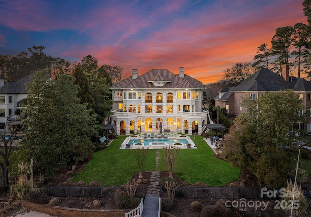 back of house at dusk with a lawn, a chimney, a balcony, and an outdoor pool