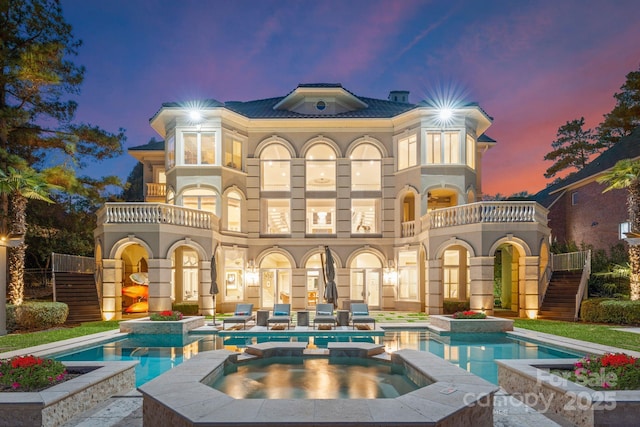 back of property at dusk with stairs, a pool with connected hot tub, a patio area, and a balcony