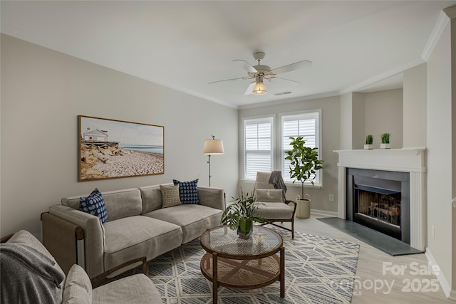 living area featuring a fireplace with raised hearth, ceiling fan, visible vents, baseboards, and ornamental molding