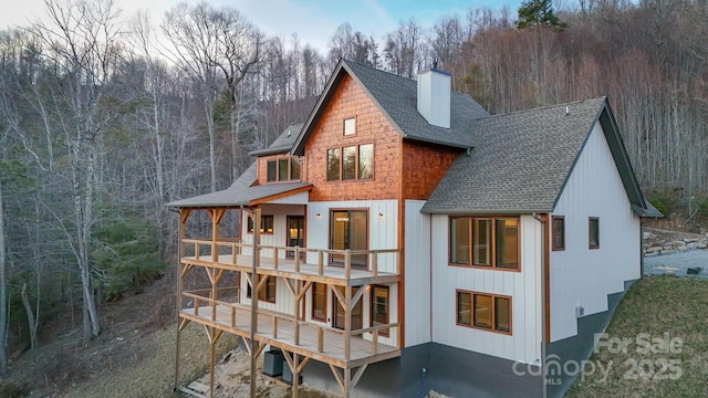 back of property with a shingled roof, a balcony, a forest view, a chimney, and central AC