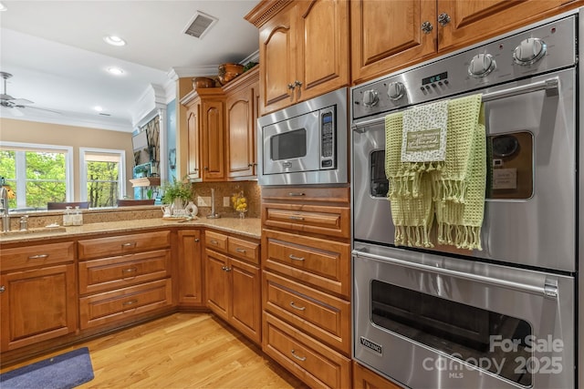 kitchen with appliances with stainless steel finishes, brown cabinetry, ornamental molding, a sink, and light stone countertops