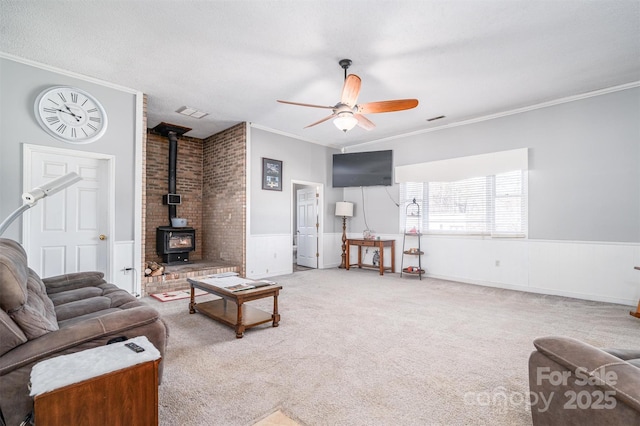 living room with carpet floors, a wainscoted wall, ornamental molding, a ceiling fan, and a wood stove