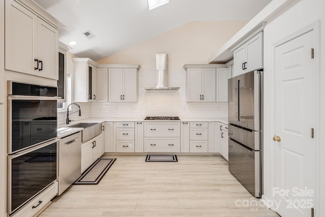 kitchen with stainless steel appliances, light countertops, vaulted ceiling, a sink, and wall chimney exhaust hood