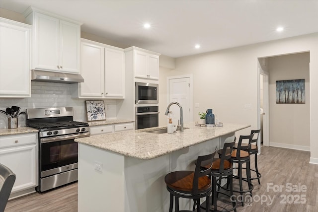 kitchen with a kitchen island with sink, stainless steel appliances, under cabinet range hood, white cabinetry, and a sink