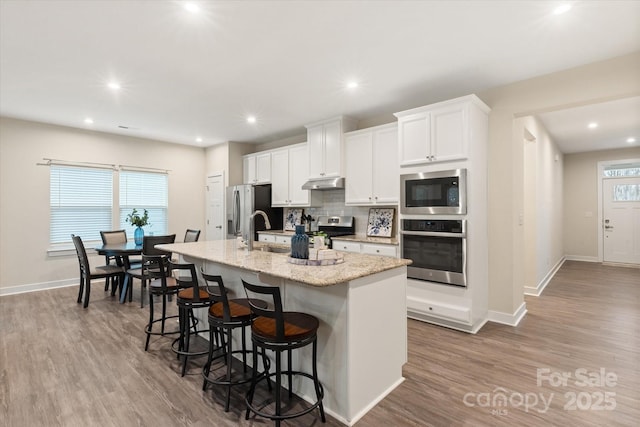 kitchen featuring white cabinets, an island with sink, appliances with stainless steel finishes, a breakfast bar, and under cabinet range hood