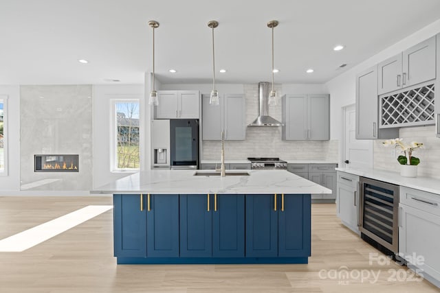kitchen featuring beverage cooler, an island with sink, wall chimney exhaust hood, a sink, and stainless steel appliances