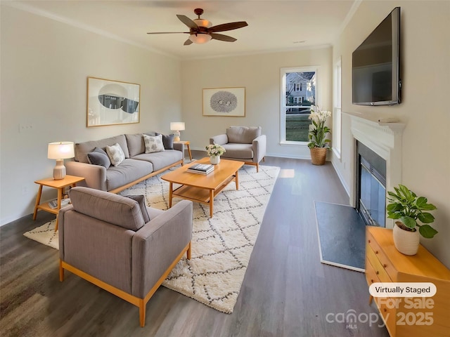 living area with ceiling fan, a glass covered fireplace, dark wood finished floors, and crown molding