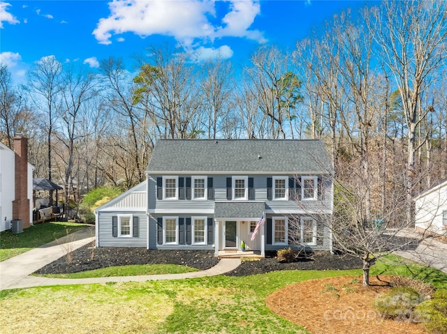 colonial-style house with driveway, central air condition unit, a shingled roof, and a front yard