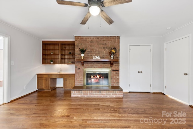 unfurnished living room with baseboards, crown molding, built in desk, wood finished floors, and a fireplace