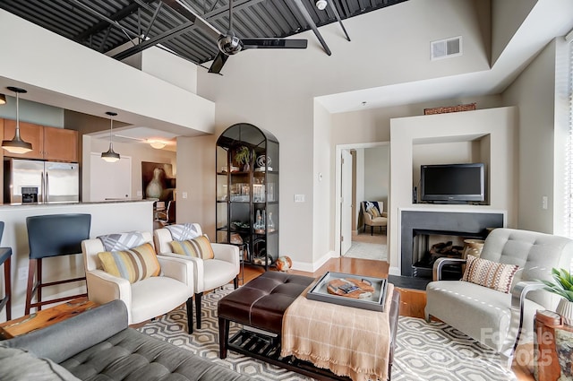 living area featuring visible vents, a fireplace with raised hearth, a towering ceiling, ceiling fan, and light wood-type flooring