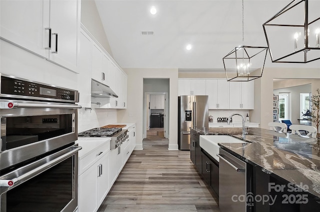 kitchen with decorative light fixtures, appliances with stainless steel finishes, white cabinets, dark stone countertops, and under cabinet range hood