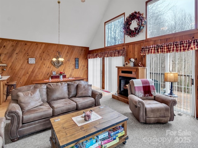 living area featuring a chandelier, light colored carpet, wooden walls, high vaulted ceiling, and a lit fireplace