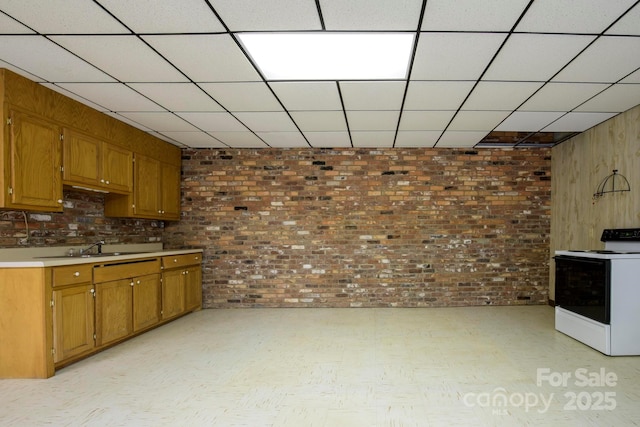 kitchen featuring a drop ceiling, brick wall, light countertops, white range with electric stovetop, and brown cabinets